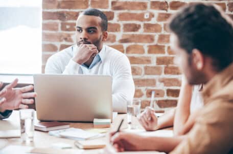 Listening to new business strategy. Thoughtful young African man holding hand on chin and listening while his coworkers discussing something sitting at the office table